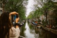 A woman taking a photo of a canal with trees and buildings on either side.