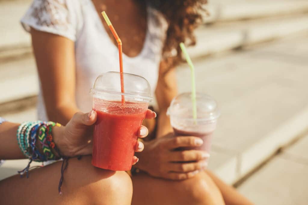 Brightly coloured smoothies in plastic cups with straws, held by a woman sitting outdoors.