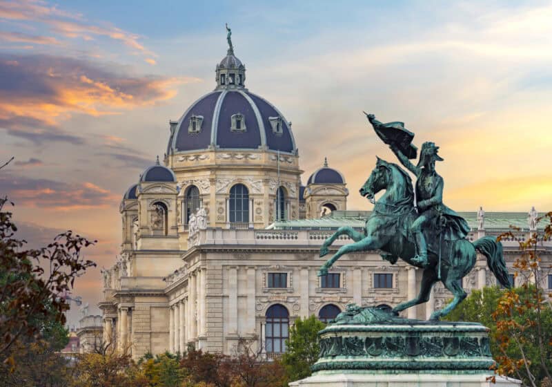 A historic building with a domed roof and an equestrian statue in front, under a colourful sky.
