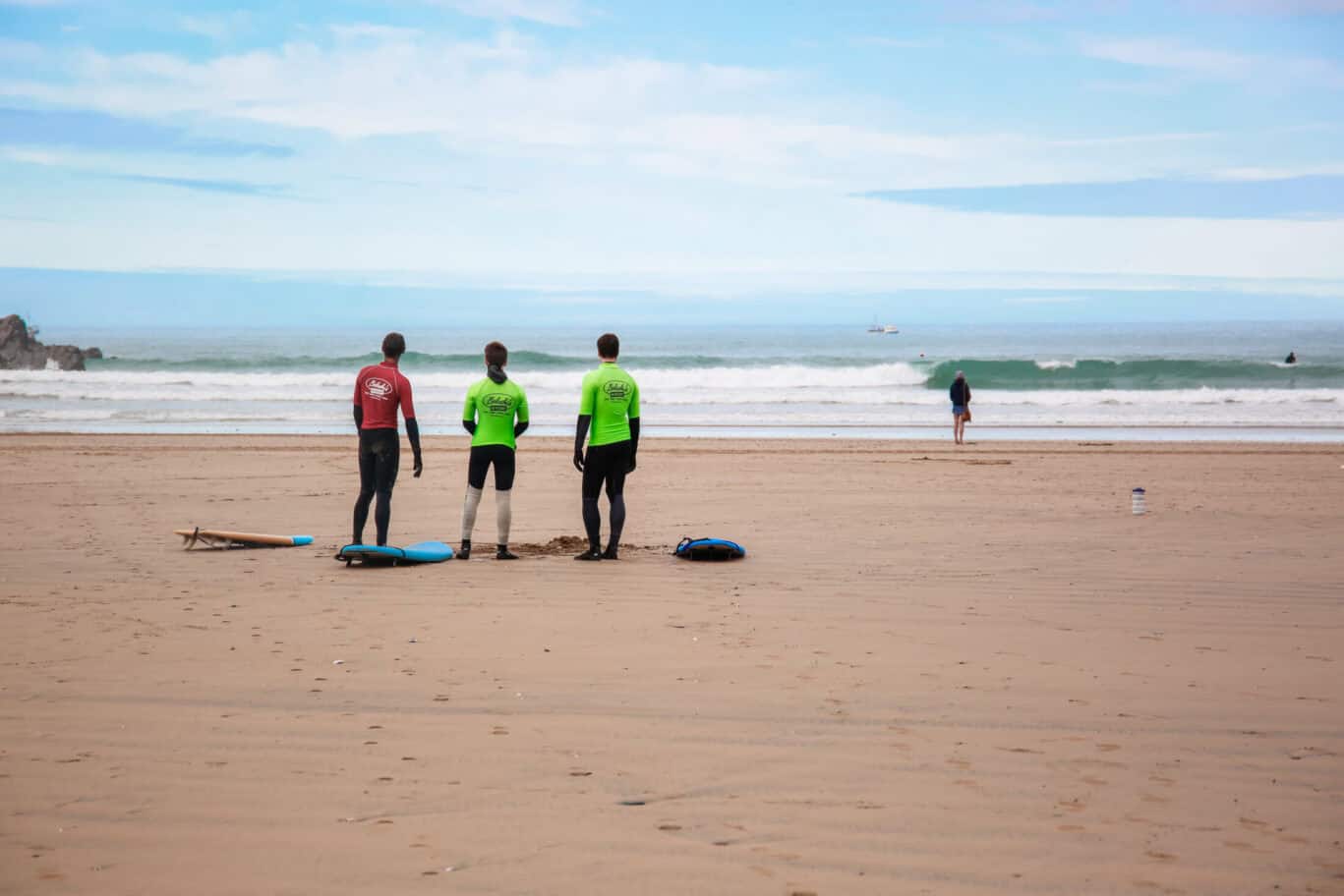 Three surfers in wetsuits with surfboards on a sandy beach, ocean waves in the background.
