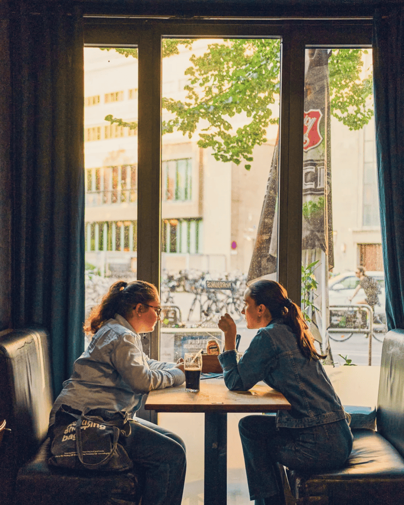 Two women chatting at a table in a social hostel cafe with large windows and street view.