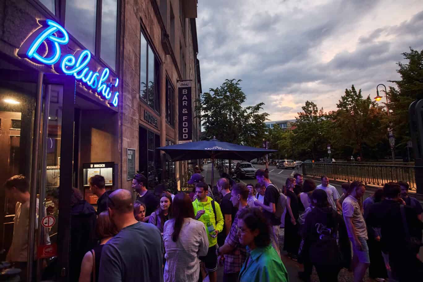 A busy social hostel bar with a neon Belushi's sign and a crowd of people outside in the evening.