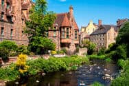 A historic building with a steep roof and bay windows beside a river with lush greenery.