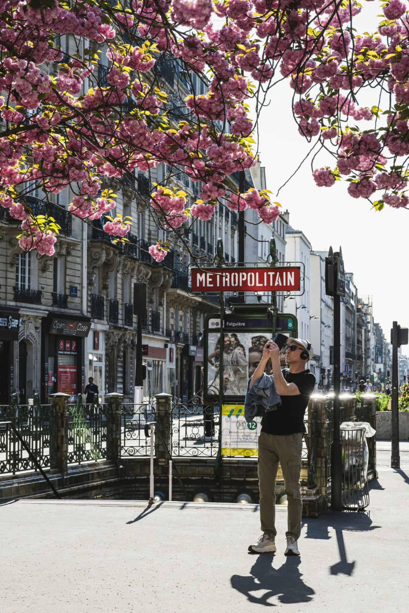 A busy city street scene with a metro station sign and a person taking a photo under blooming cherry.