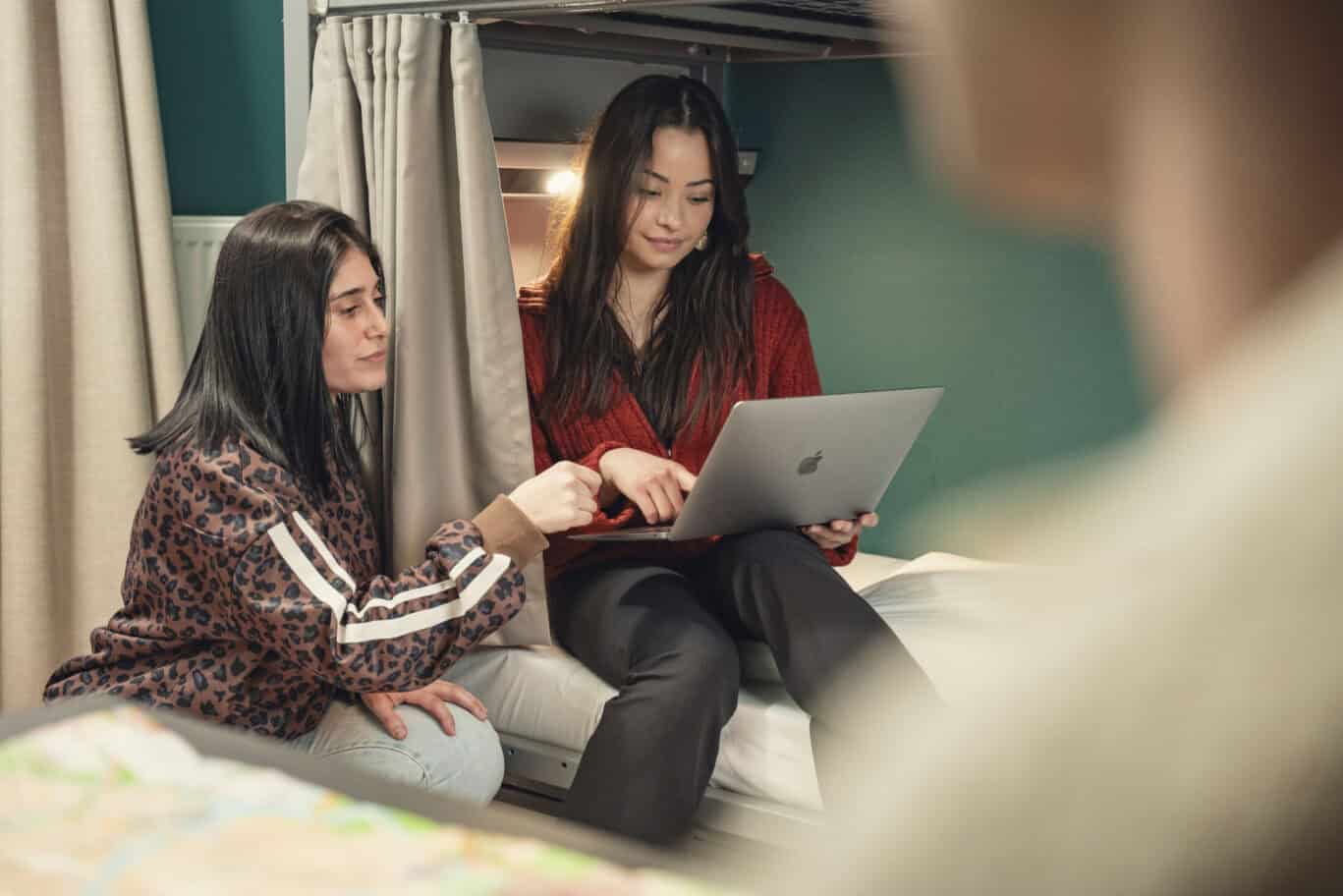 Two young women studying together on a laptop in a dorm room setting.
