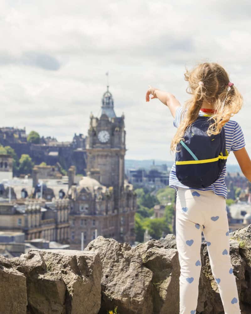 A young girl with a backpack pointing at a city clock tower from a rocky viewpoint.