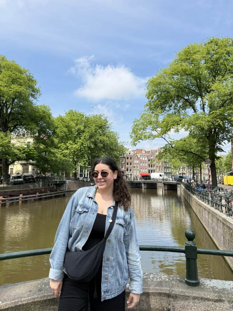 A woman standing on a bridge over a canal with trees and buildings in the background.