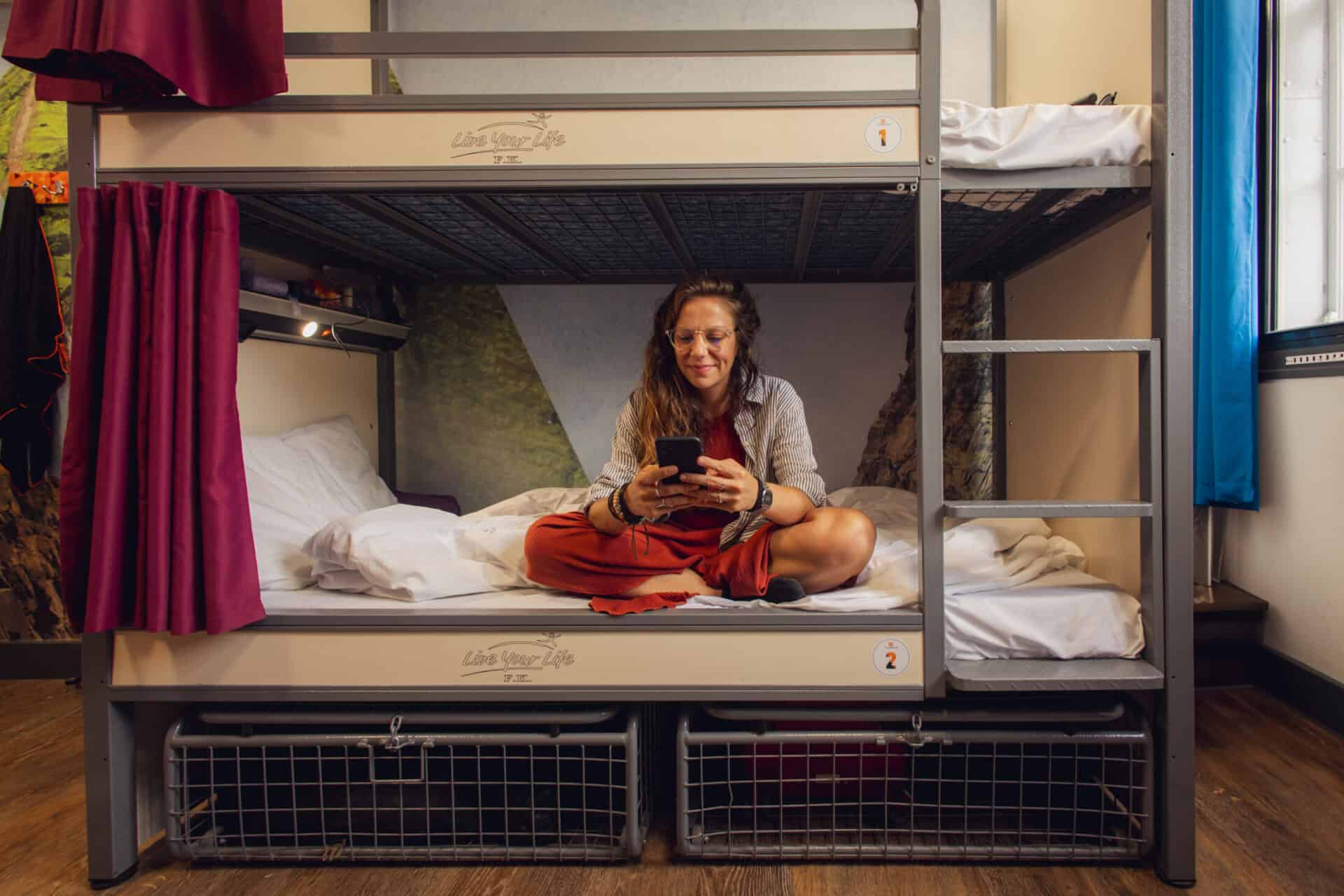 A traveller sitting on a bunk bed in a hostel dorm room, using a smartphone.