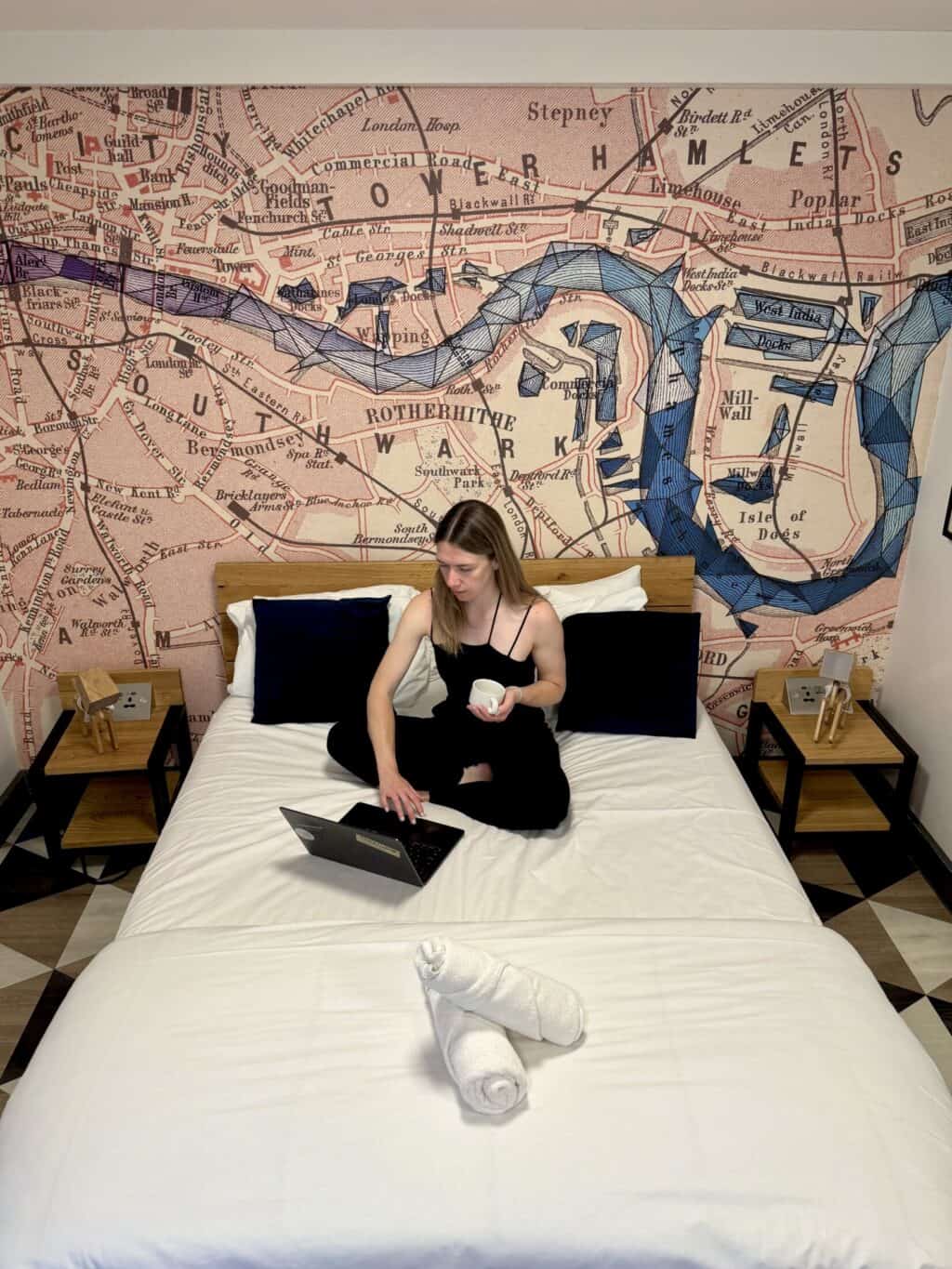 A woman working on a laptop on a queen bed in a hotel room with a city map mural.