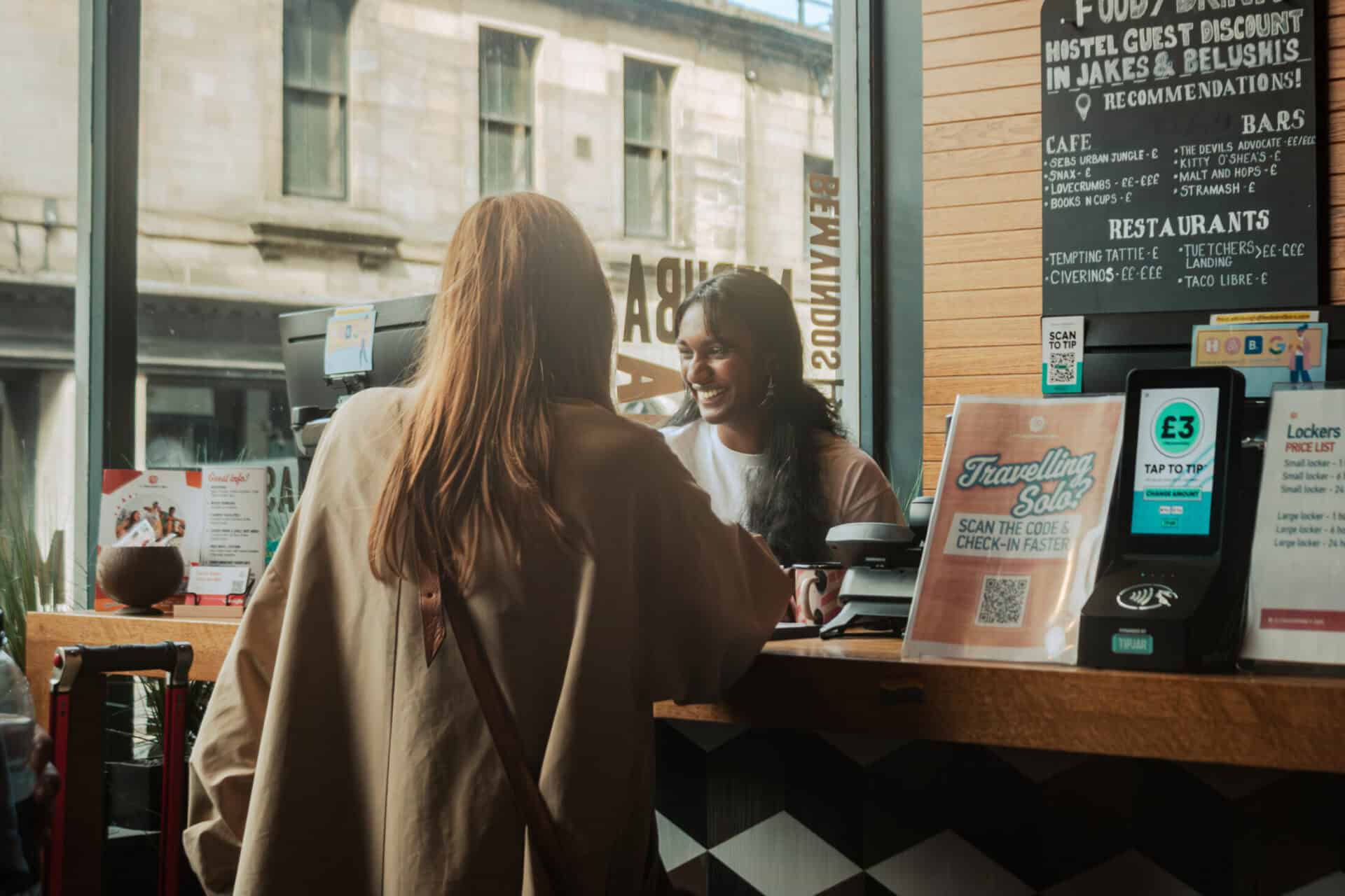 A social hostel bar scene with two women at the counter, one ordering and the other serving, in a St.