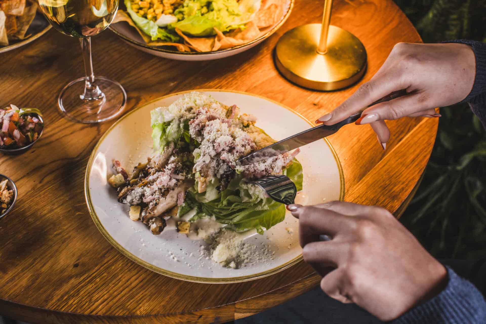 A person cutting into a salad on a wooden table with wine glasses and a candle.