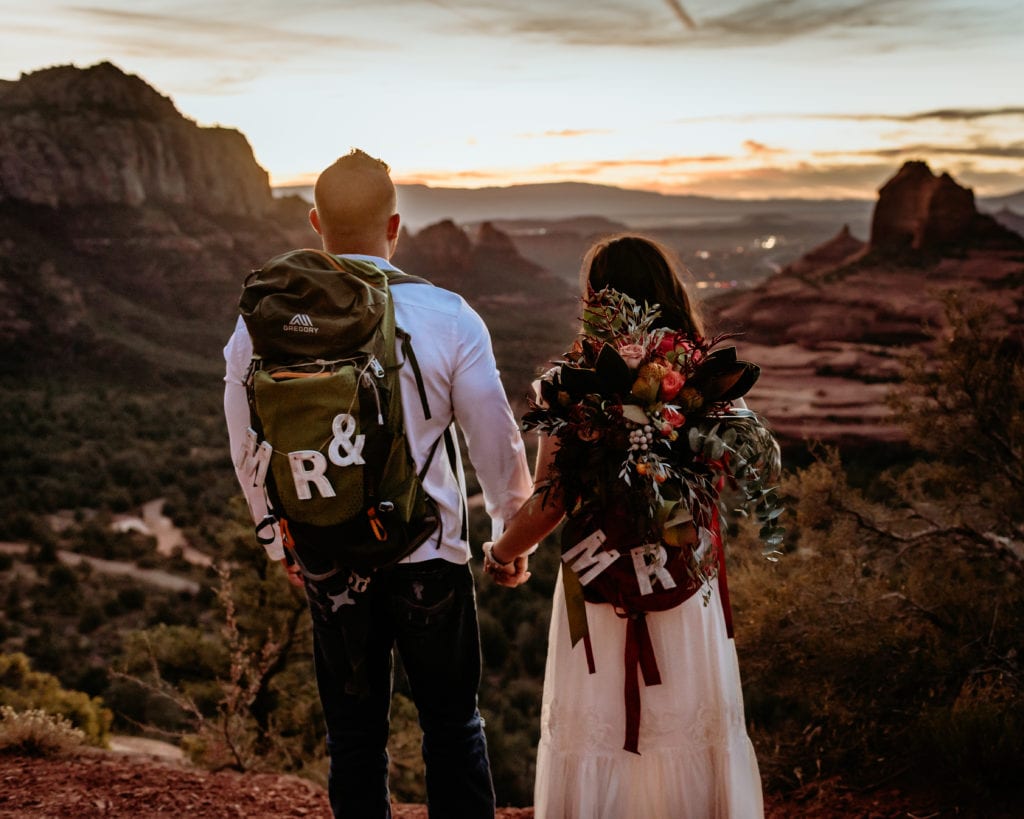 Elopement couple holding hands with their backpacks overlooking Sedona