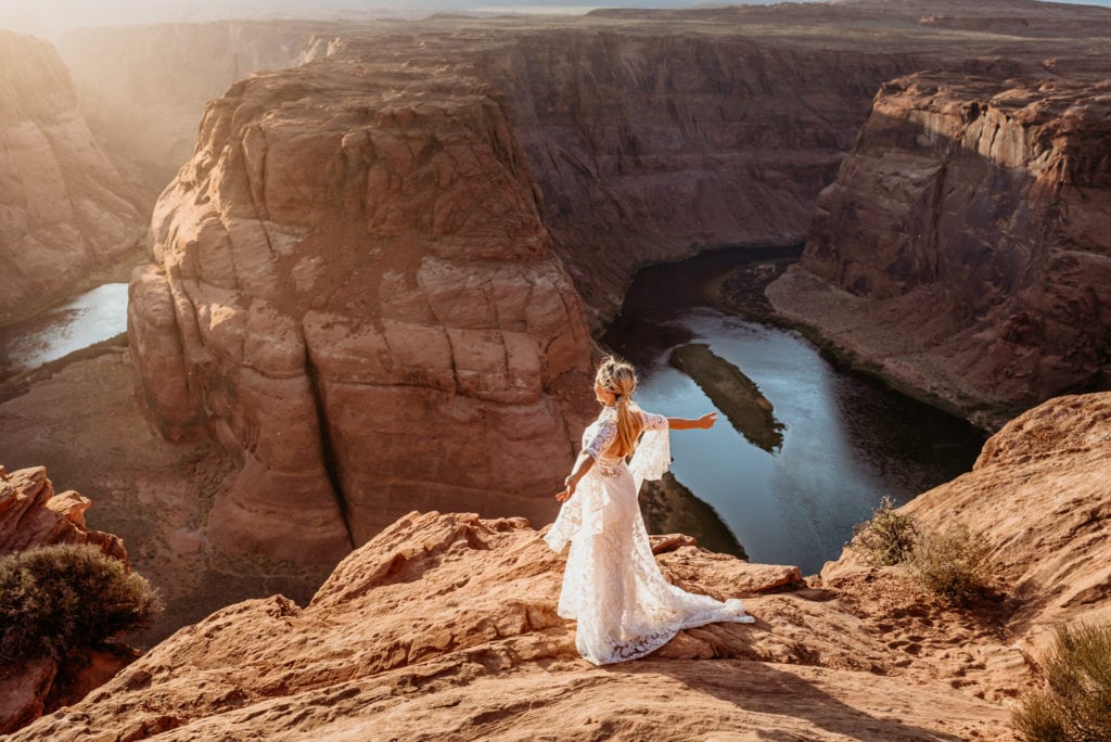 Bride standing arms outstretched as the wind blows and sun shines against her face at the edge of Horseshoe Bend in Page, Arizona