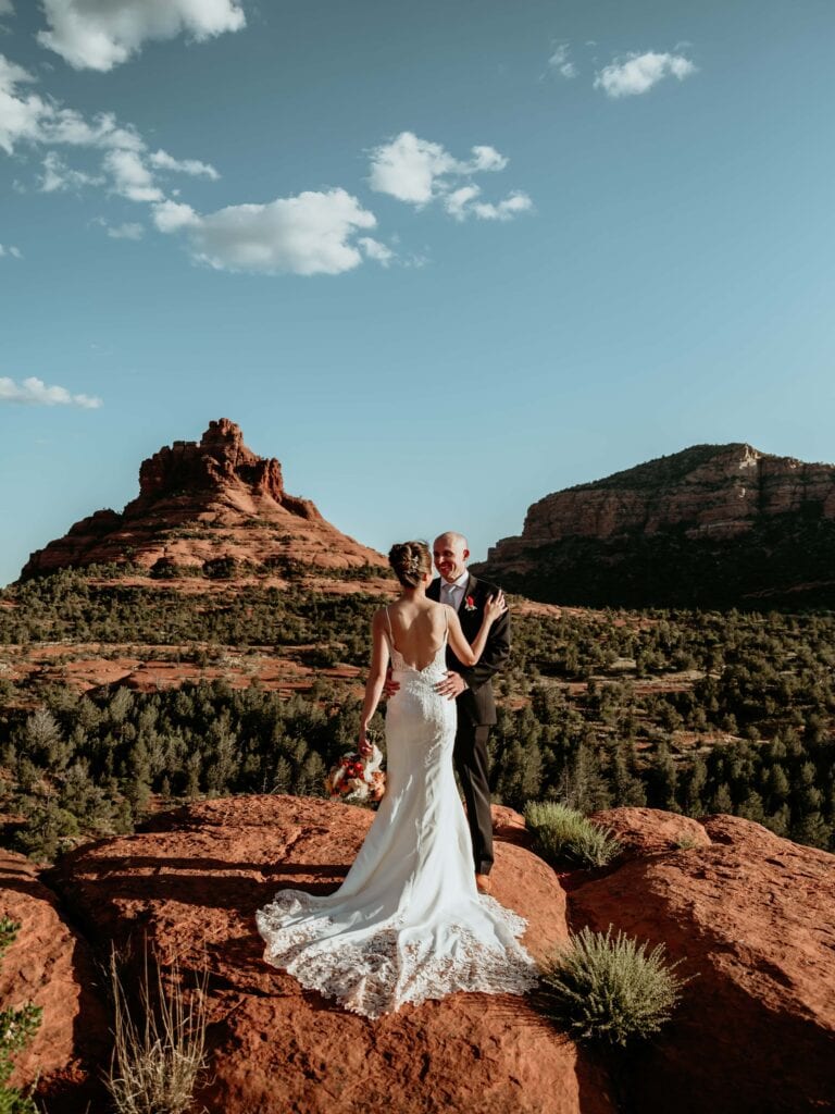 Couple embraces with the train of the bride's dress as they stand with blue skies and Bell Rock in the background