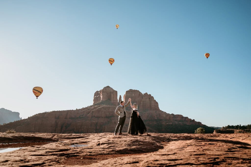 Bride in black dress dancing with groom with hot air balloons rising behind them in Sedona