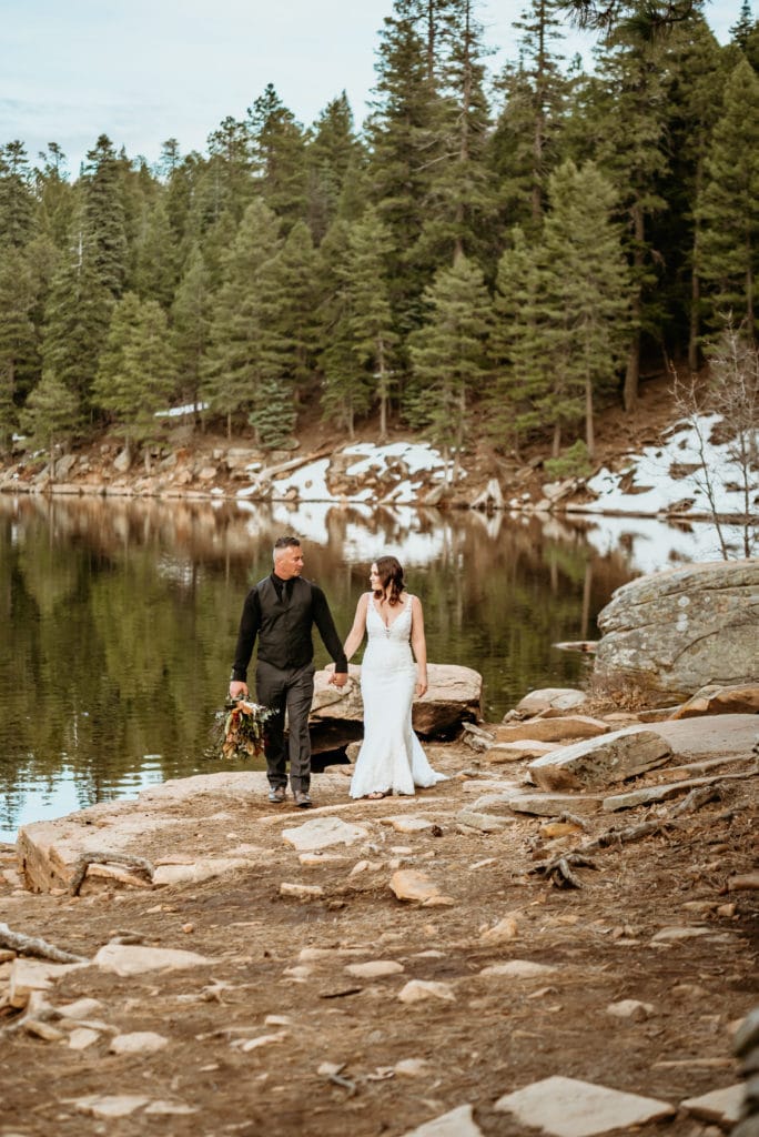 Bride and groom walking hand in hand next to winter lake lined by pine trees and patches of snow