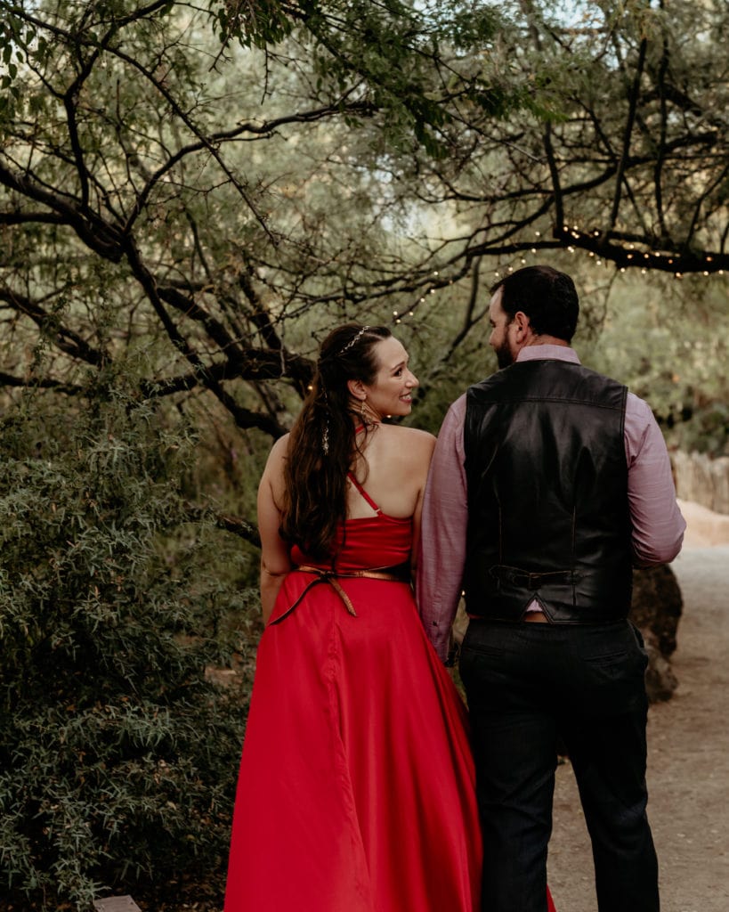 Bride looking over her shoulder as she and groom walk through tree lined trail in her vibrant red wedding dress