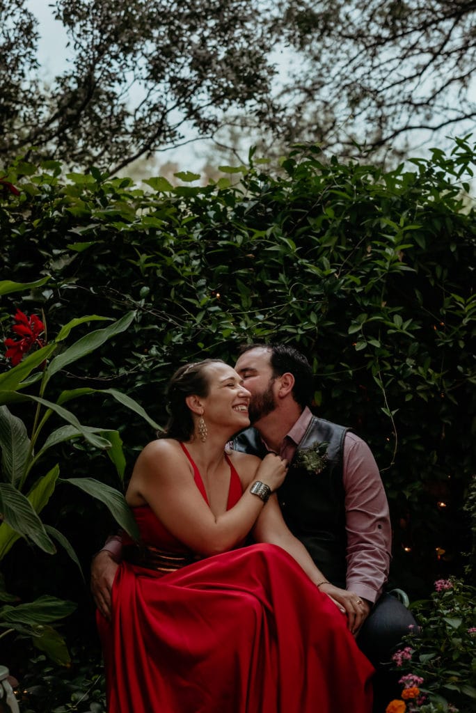 Bride n bright red dress laughing as groom whispers in her ear surrounded by dark green vines at botanical gardens