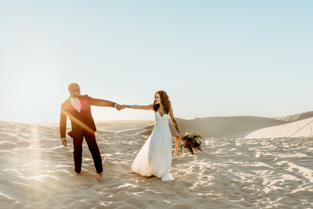 Elopement couple playfully walk hand in hand, barefoot, across the endless desert sand dunes