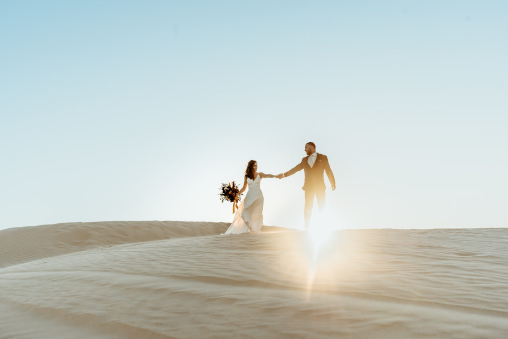 Elopement couple walking hand in hand down Glamis Sand Dunes as the sun sets behind them