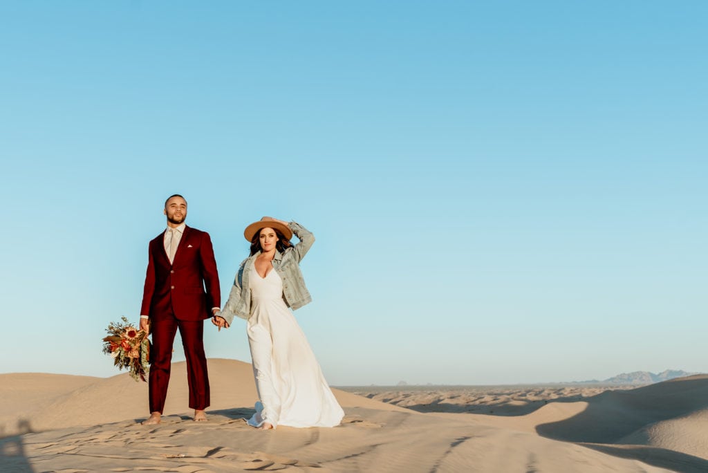 Bride in boho denim jacket and brimmed hat, groom in dark maroon suit, both barefoot standing on top of sea of sand