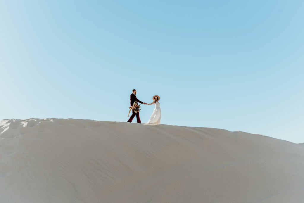 Groom carrying bouquet as bride leads him across the sand during their minimalistic desert elopement