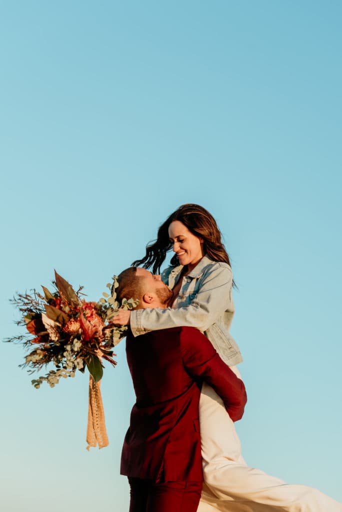 Bride smiling down on groom with her deep, vibrant bouquet as he lifts her into the sunset
