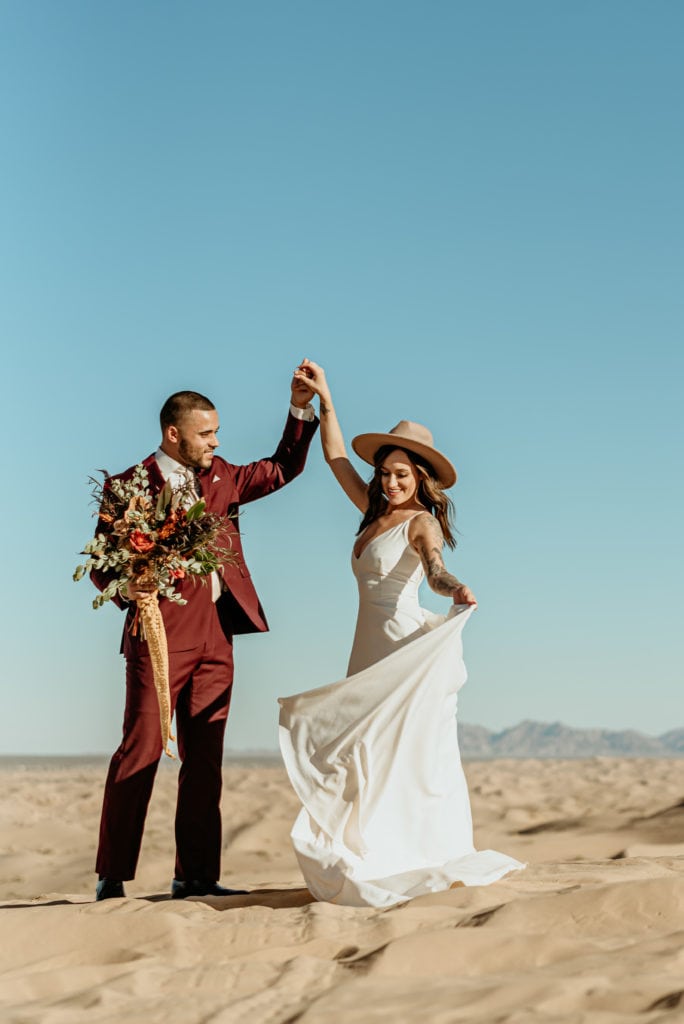 Groom in maroon tux spins his bride in a long minimalist white dress and boho hat
