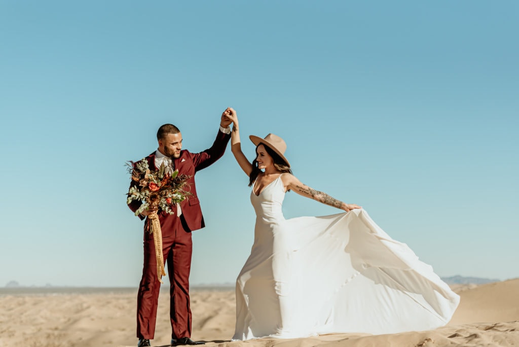 Elopement couple dancing as the bride's dress flows in the wind sweeping across the Imperial Sand Dunes