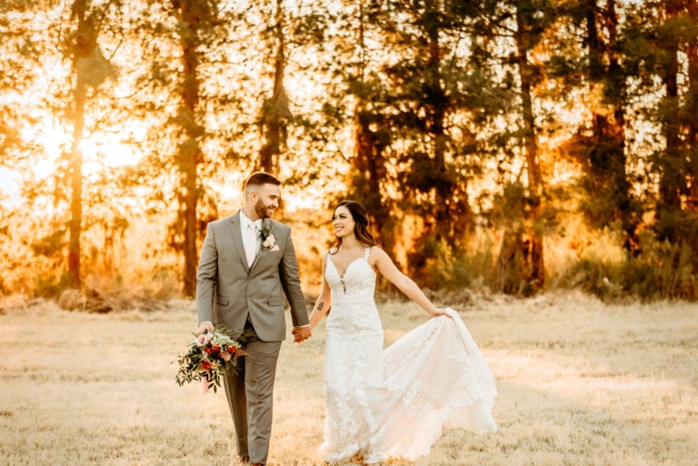 Bride and groom walking hand in hand while golden rays shine through a wall of evergreens