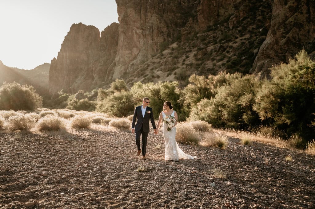 Couple walking hand in hand with the Bulldog Cliffs at the Salt River towering behind them during their Arizona elopement by Stratus Photography