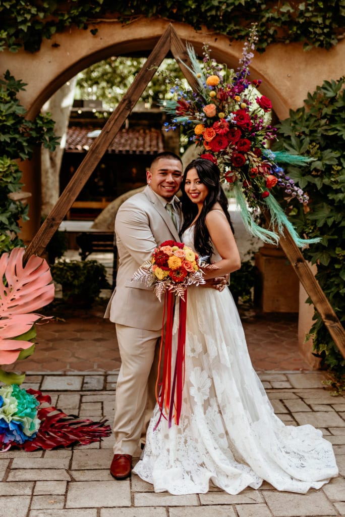 Bide and groom in front of wedding arch decorated with vibrant flowers