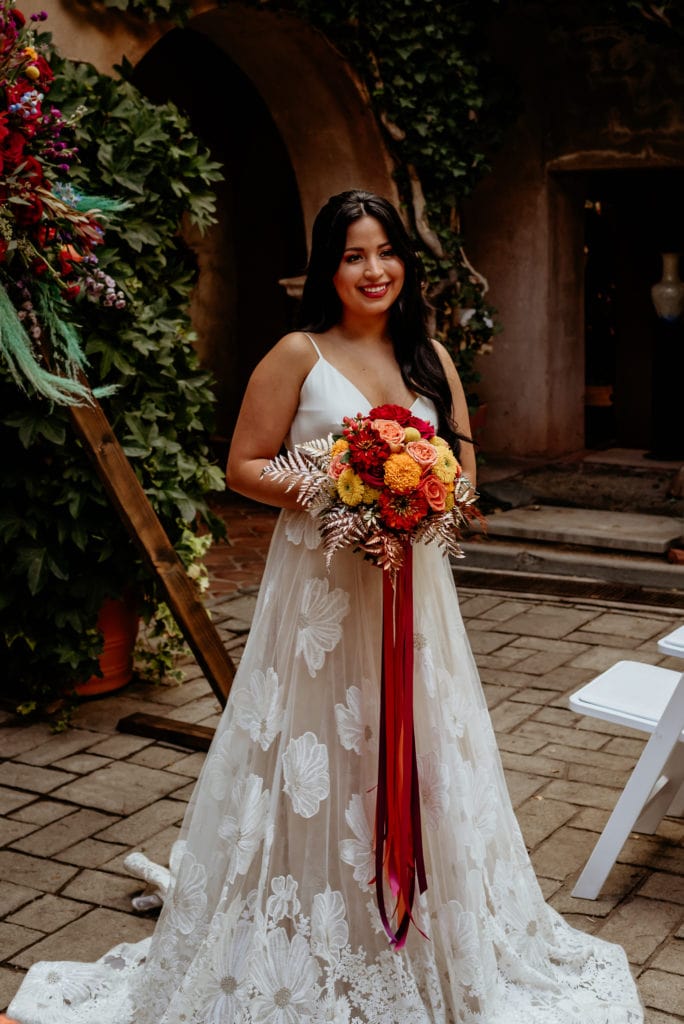 Bride in long floral lace dress holding bouquet with yellows, reds, orange and gold touches