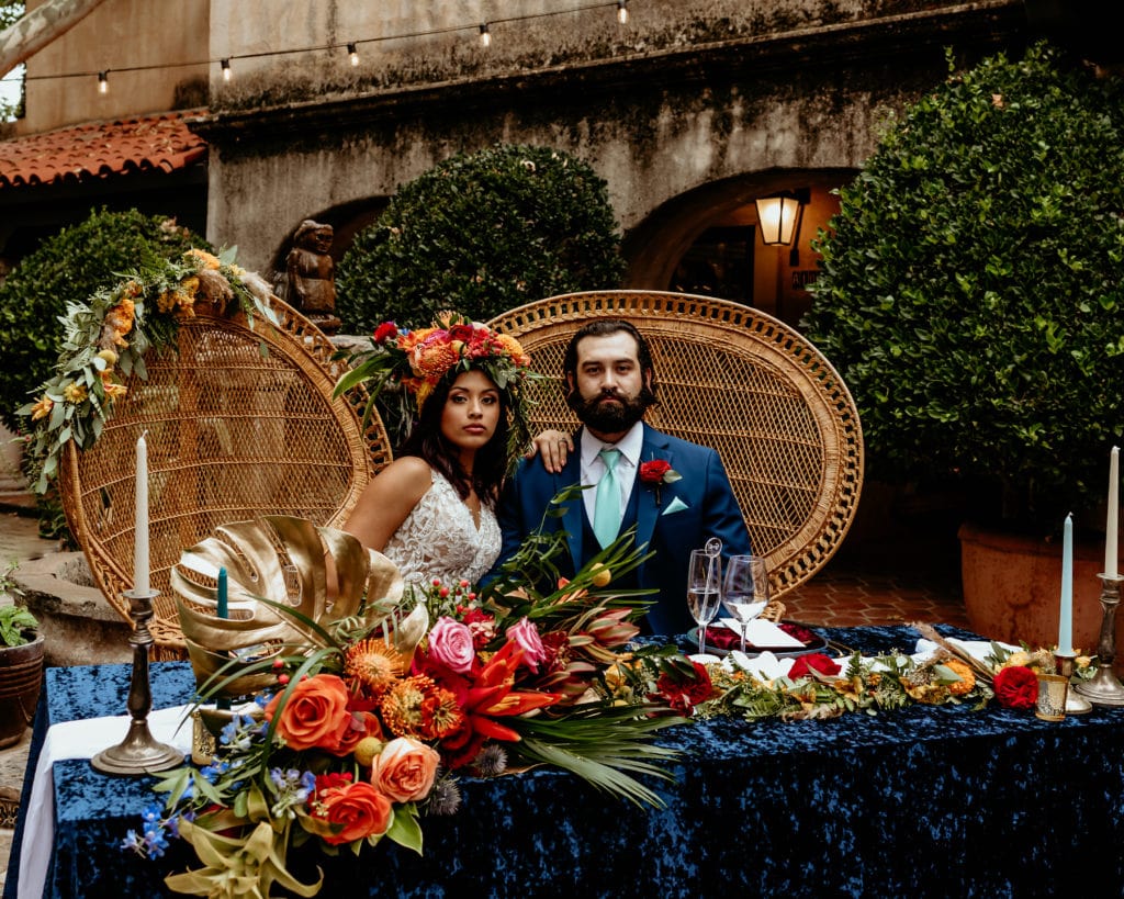 Bride and groom looking stoic and regal in wicker peacock chairs with tropical headdress and vibrant florals during their Tlaquepaque wedding
