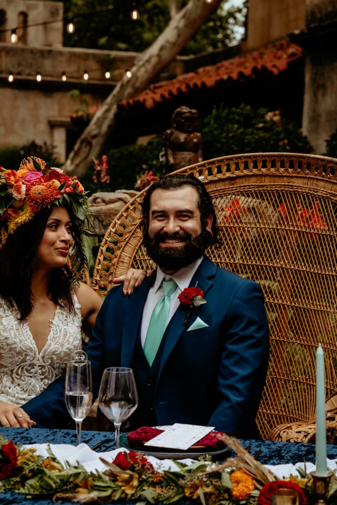Groom laughing as bride with tropical headdress looks at him lovingly in courtyard wedding