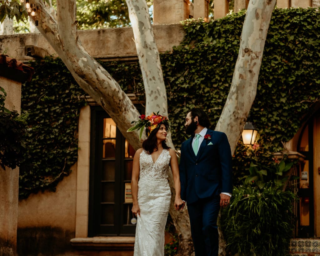 Bride in long lace gown as she gazes at her husband in the courtyard