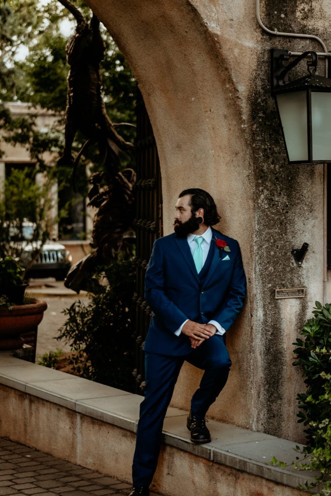 Groom in navy blue suit and teal tie leaning against stucco wall