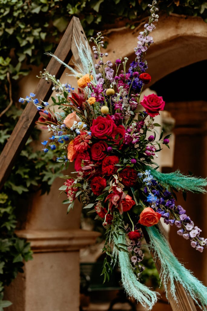 Gorgeous ceremony arch with deep reds, bright purple, orange, yellows, and sprigs of teal