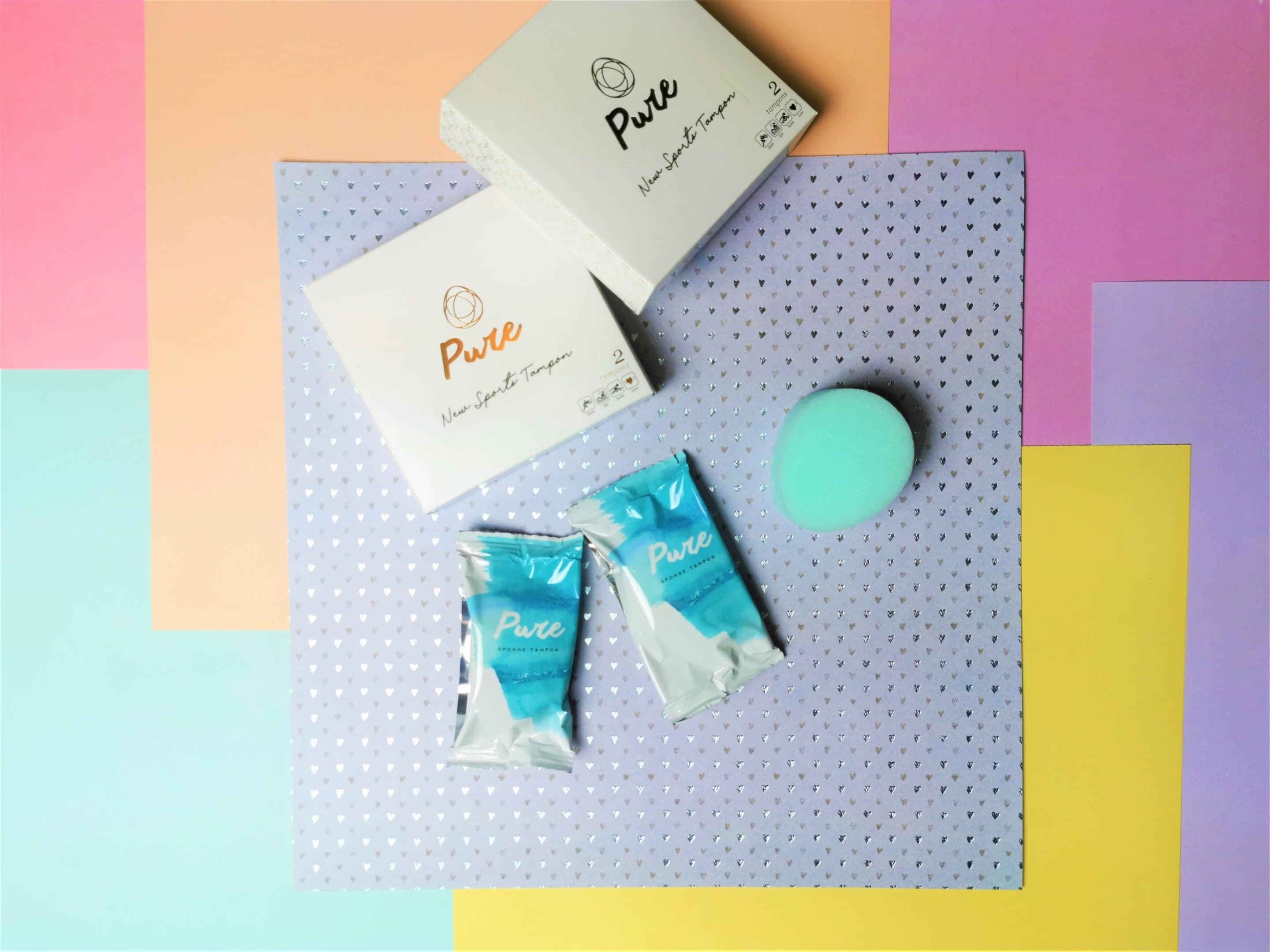 Twee verpakkingen van beppy pure, witte doosjes met in het goud de naam er op. Daarvoor liggen twee beppy tampons, per stuk verpakt en daarnaast ligt een uitgepakte Beppy Pure tampon. Het is een ovaal, blauw sponsje.