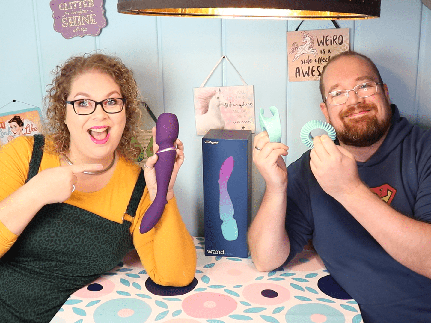 Tess en Jay zitten aan de keukentafel. Tussen hun in staat de verpakking van de we-vibe wand. Tess heeft de wand vast en Jay de twee opzetstukken van de We-Vibe Wand.