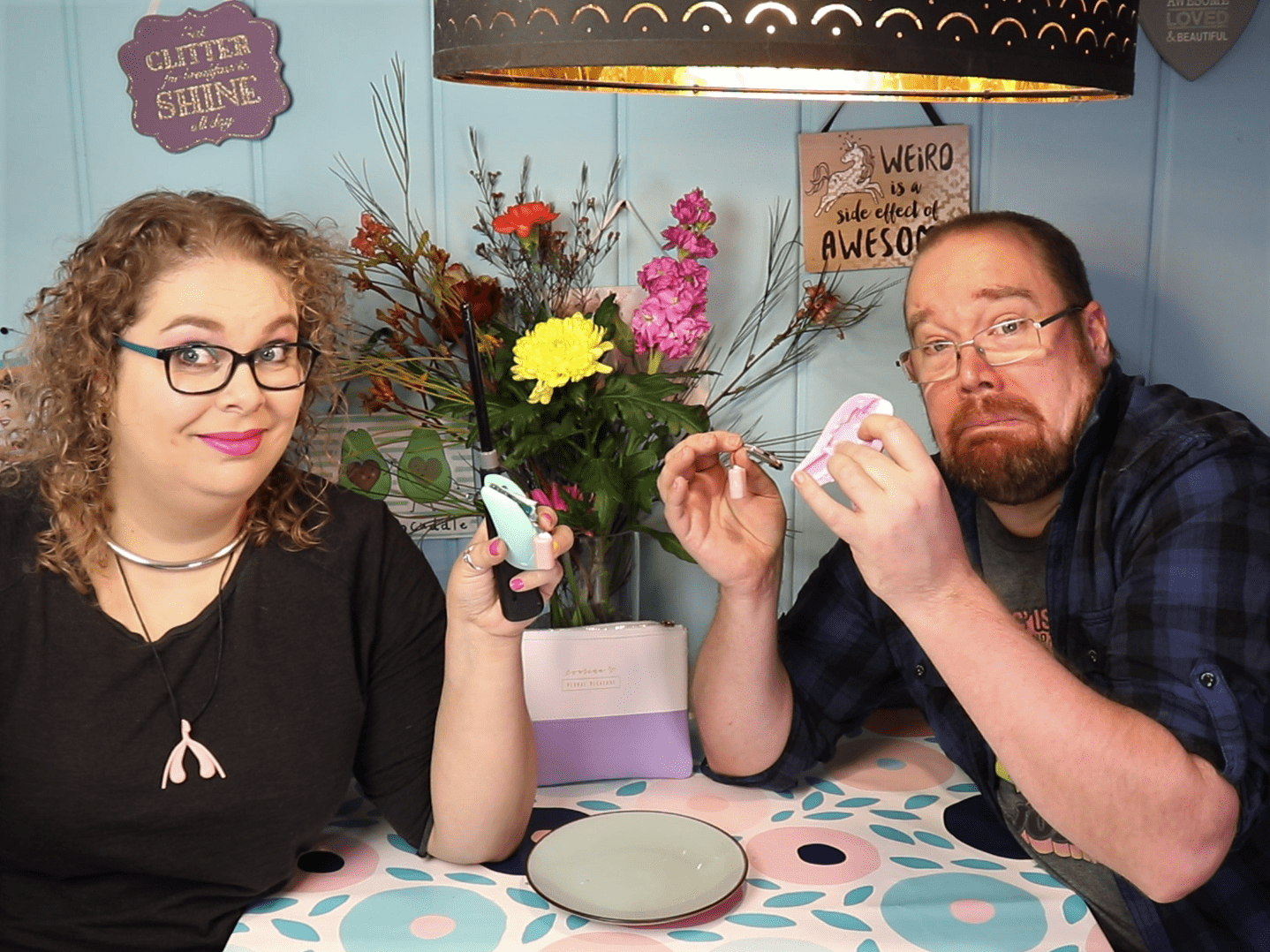 Tess en Jay zitten aan de keukentafel. Tess heeft de L'Eve vast, maar de motor hangt er uit. Ze heeft ook een aansteker vast en ze kijkt een beetje moeilijk. Jay heeft de AliExpress toy vast en in zijn andere hand de motor en de batterij. Hij kijkt geschrokken.