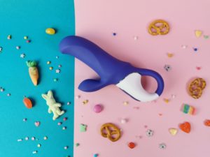 De satisfyer vibes magic bunny ligt op een petrolkleurig en lichtroze achtergrondje, met allerlei gekleurde sprinkles. De vibrator is een duovibrator, hij is paars en het handvat is deels wit.