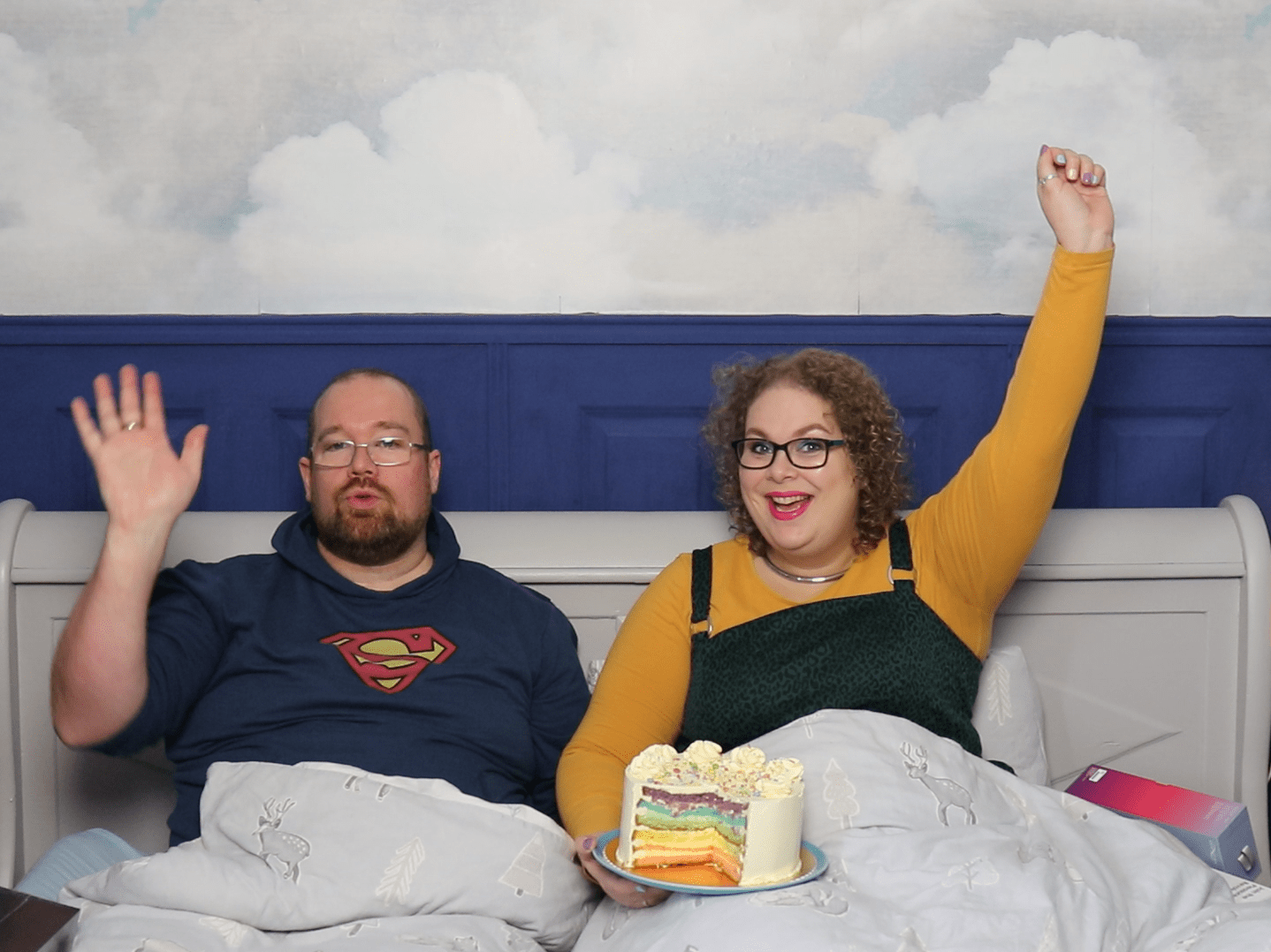 Tess en Jay zitten in hun bed. Tess heeft een grote regenboogtaart op haar schoot en steekt haar andere arm in de lucht. Jay steekt een arm ook een beetje in de lucht.