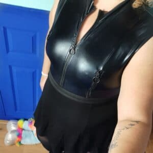 Tess staat naast het bed en draagt de lovehoney fierce wet look body met daar over een zwart rokje.