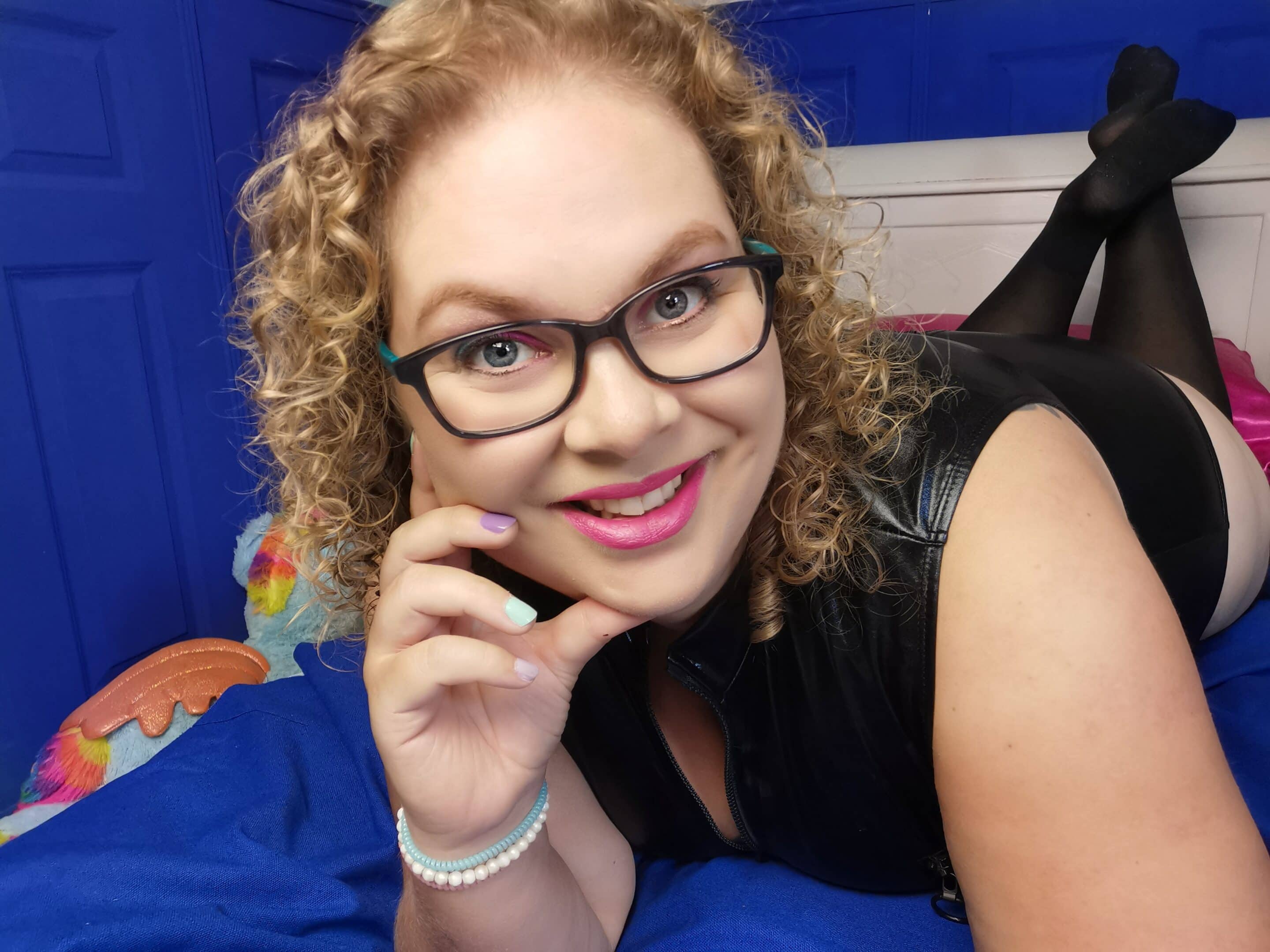 Tess ligt op bed, op haar buik, en ze draagt de Lovehoney Fierce Wet Look Body en kousen.