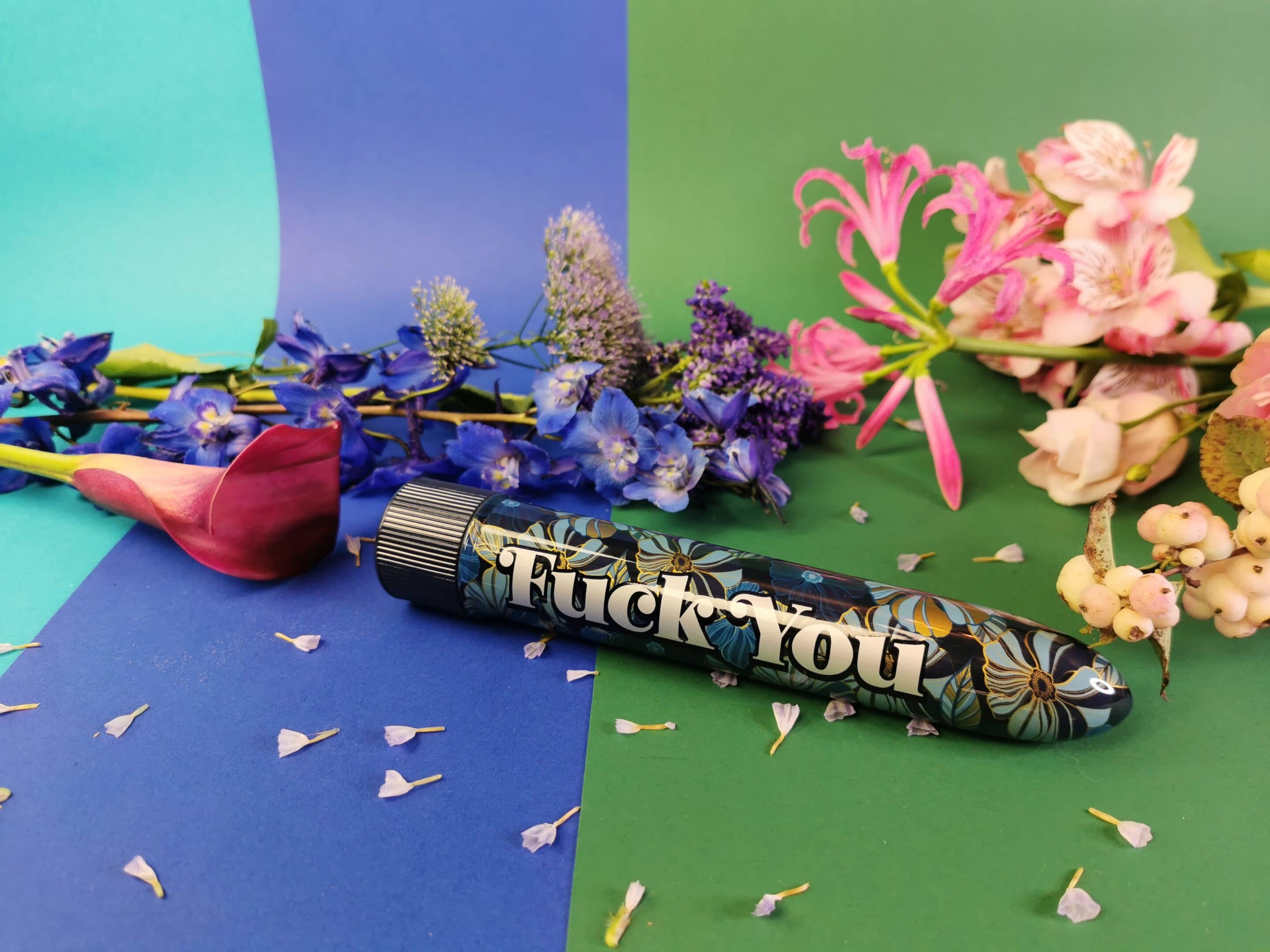 De Naughty Bits Fuck You Vibrator ligt op een blauw en groen ondergrondje, tussen allemaal bloemen en bloemblaadjes. De vibrator zelf heeft een bloemenprintje, vooral in goud en verschillende tinten blauw. En er staat vrij groot de tekst 'fuck you' op.
