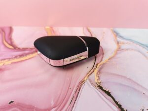 De satisfyer love triangle is een zwarte, driehoekige toy met een gouden randje. Op het randje zie je de merknaam satisfyer staan. Het tuutje is afgesloten met een dopje. Het geheel ligt op een roze met goud ondergrondje.