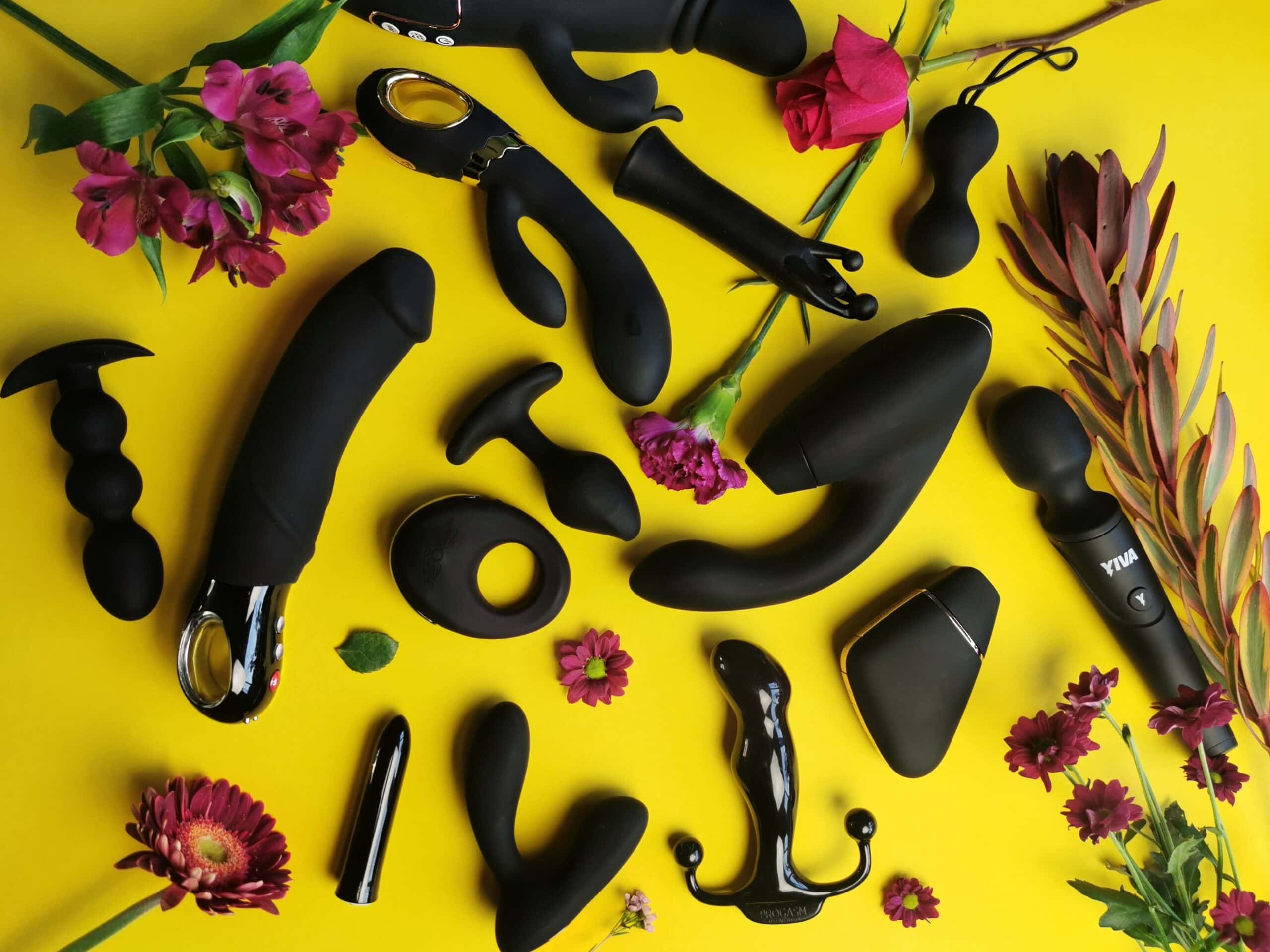 Een aantal zwarte seksspeeltjes liggen op een geel ondergrondje. Er tussen liggen wat bloemen.