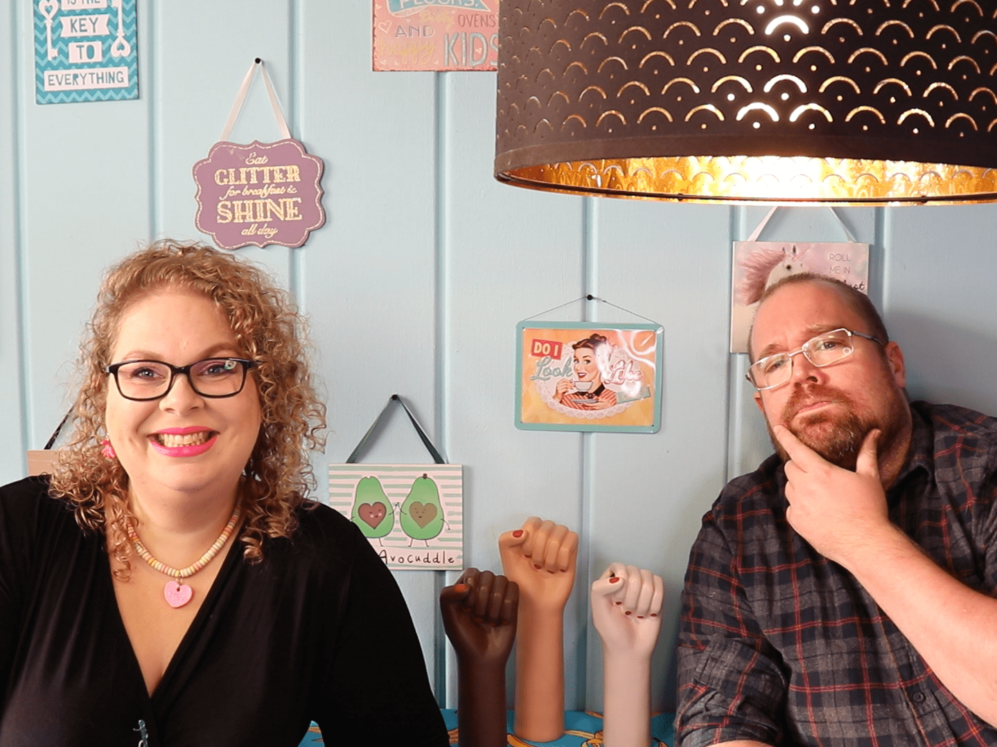 Tess en Jay zitten aan de keukentafel. Tess lacht en Jay kijkt bedenkelijk met een vinger onder zijn kin.