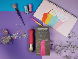 De fun factory sundaze naast de verpakking waarvan je de achterkant ziet. De achterkant is donekrbruin met een shiny foto van het bovenste deel vand e toy en wat tekst.